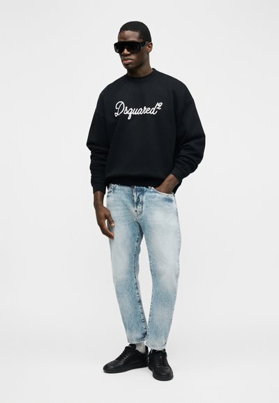 Άντρας που φοράει μαύρο φούτερ Dsquared2, ανοιχτόχρωμο μπλε τζιν, μαύρα αθλητικά παπούτσια και μεγάλα μαύρα γυαλιά ηλίου, στέκεται με το ένα χέρι στην τσέπη.