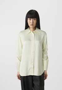 Blusa in satin gialla chiara con colletto a punta, chiusura frontale con bottoni e maniche lunghe. Texture liscia; vestibilità leggermente ampia.