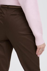 Tuvplāns cilvēkam, kurš valkā brūnas bikses ar melnu jostu un gaiši rozā ar garām piedurknēm džemperi, roka atbalstīta uz augšstilba.