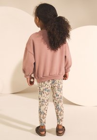 Roze sweatshirt met losse mouwen, bloemenleggings met patroon in crème, blauw en bruin, en bruine sandalen. Achteraanzicht tegen een neutrale achtergrond.