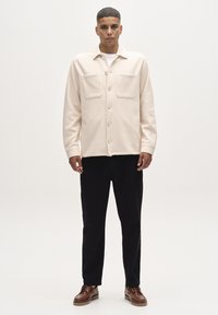 Beige nööbiga särk kahel rindkere taskulisega, kantud valge särgi peal. Mustad püksid ja pruunid kingad täiendavad riietust. Lihtne ja puhas disain.