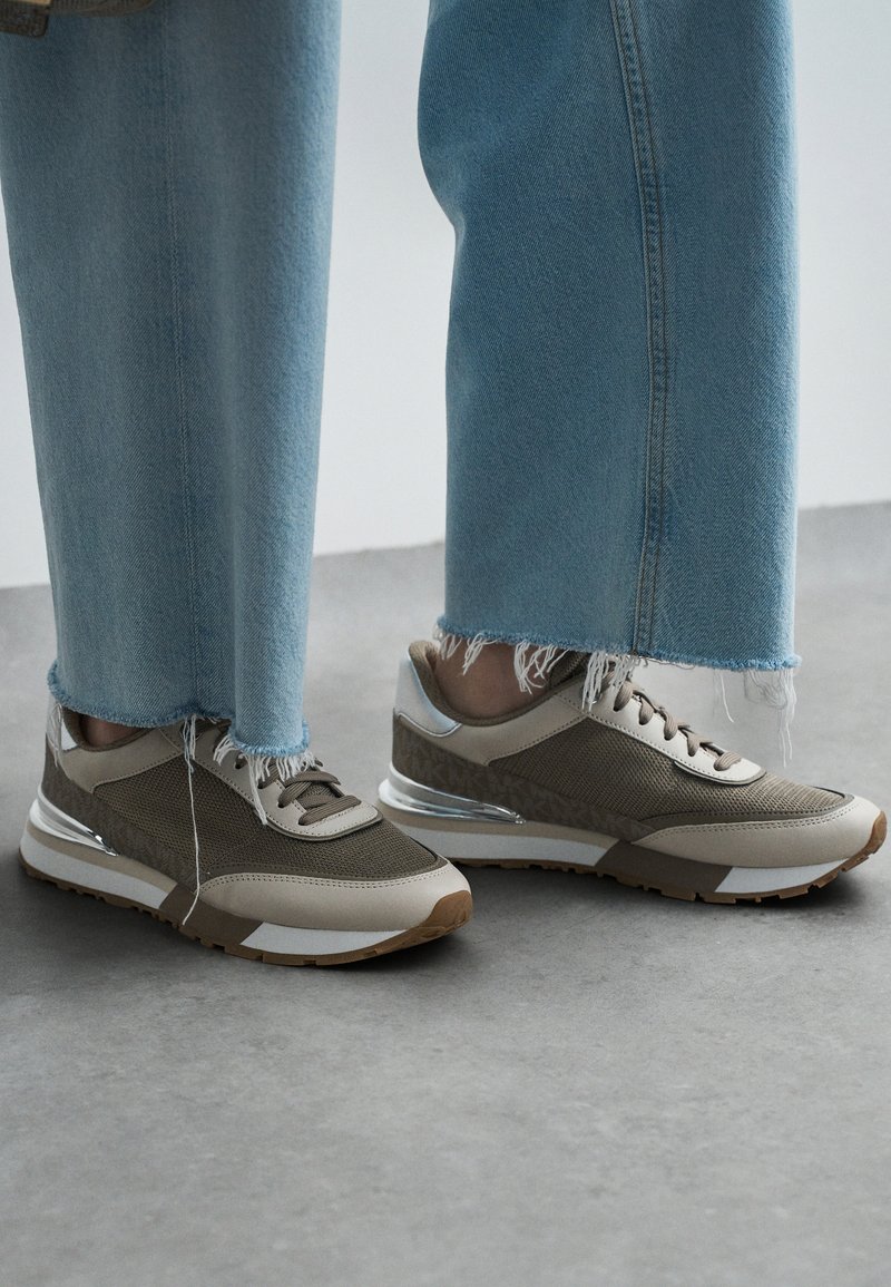 Sneakers leggeri con tomaia in rete di colore oliva e beige, un motivo testurizzato, suola beige e dettagli riflettenti. Indossati con jeans sfrangiati.