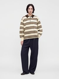 Personne aux cheveux courts et foncés porte une chemise rayée beige et marron à manches longues, un pantalon noir ample et des chaussures noires, debout devant un fond blanc.