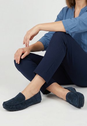 Osoba sedící se zkříženýma nohama, oblečená v tmavě modrých kalhotách, modré košili na knoflíky a tmavě modrých semišových mokasínách.