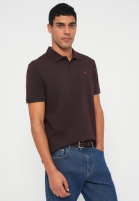 Burgundská polokošile z texturované látky, s krátkými rukávy a límečkem, s malým červeným logem na levé straně hrudi. Nošena s modrými džínami.