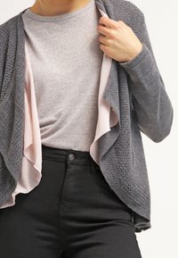 Cardigan grigio lavorato a maglia indossato sopra una camicia grigia chiara con foderatura interna rosa pallido, abbinato a pantaloni neri; presenta un design con scollo drappeggiato.