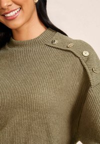 Olivza zaļas ribotas adītas džemperis ar apaļu kakla izgriezumu un dekoratīvām pogām uz pleca. Mīksta tekstūra un brīvi pieguļošs stils.