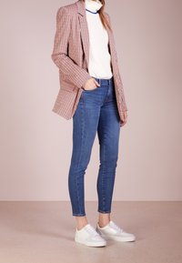 Persona con un suéter blanco de cuello alto, blazer a rayas rojas y blancas, jeans ajustados azules y zapatillas blancas, de pie con una mano en el bolsillo.