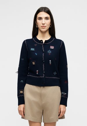 Mujer con cabello largo y oscuro que lleva un cárdigan azul marino con botones y parches bordados multicolores y pantalones cortos beige, de pie frente a un fondo gris.