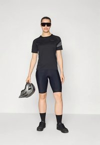 Camisola preta de ciclismo com mangas curtas, calções escuros de ciclismo e ténis pretos. Segura um capacete cinzento, usa óculos de sol. Textura de tecido suave.