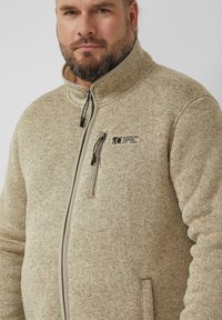 Veste en polaire beige avec une poche poitrine zippée, un col montant et une fermeture éclair grise contrastante. Présente un motif en tricot texturé.