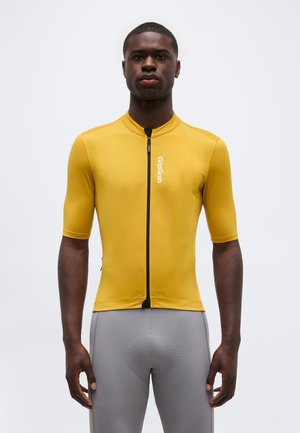 Man draagt een nauwsluitend geel wielershirt met ritssluiting en grijze gewatteerde wielershorts, staande tegen een effen achtergrond.