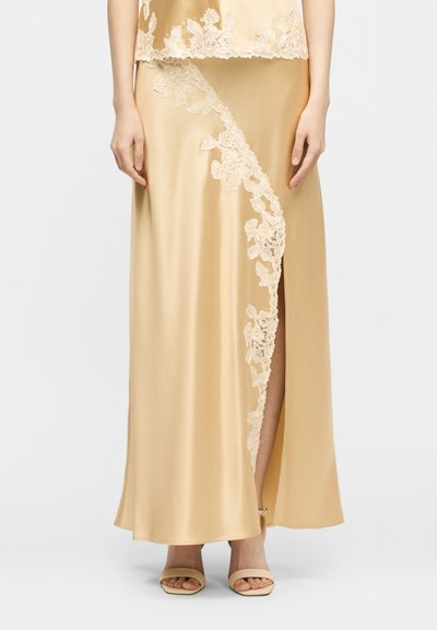 Μακριά φούστα από σατέν σαμπάνια με δαντελένια επένδυση και πλάγια σκίσιμο, συνδυασμένη με τοπ με δαντελένιο τελείωμα και μπεζ πέδιλα με ανοιχτά δάχτυλα.