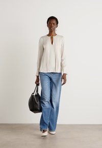 Beige langermet skjorte med detaljer i nøkkelhullet foran, sammen med lyseblå vide jeans. Modell holder en svart håndveske og har på seg beige sko.
