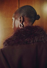 Cappotto marrone con collo in pelliccia, completo di un grande orecchino d'argento con un accento rosso. Capelli acconciati corti con un piccolo chignon.