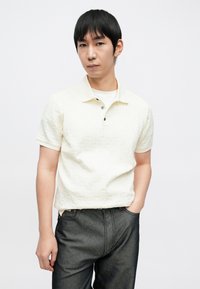 Poloshirt mit Kragen und kurzen Ärmeln in hellem Creme mit einem strukturierten geometrischen Muster, kombiniert mit dunkelgrauer Jeans aus Denim.