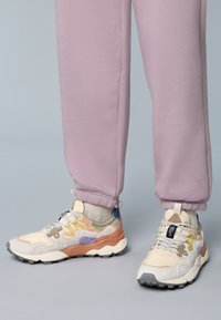 Färgglada sneakers med beige, bruna och lila detaljer, texturerade material och grå gummisulor. Parat med ljuslila joggers.