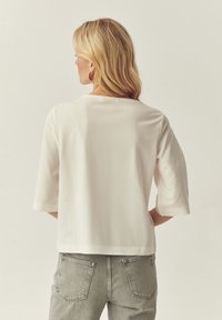 Haut blanc en coton ample avec manches trois-quarts, col rond et texture lisse, associé à un jean gris en denim.