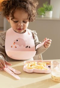 Roze siliconen slabbetje met een varkensontwerp, bijpassende bestek, en een roze bord in dierenvorm met pasta. Zachte, gestructureerde materialen, speelse accenten.