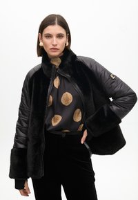 Black puffer jacket with faux fur trim, zip closure, and wide sleeves. Paired with a dark shirt featuring large gold polka dots and black pants.