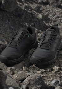 Pair of black trail running shoes with laces on rocky, uneven ground, highlighting rugged outdoor footwear design.