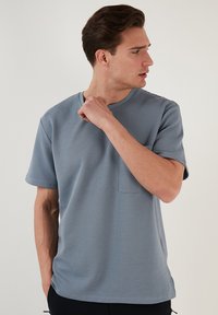 Svetle modré tričko s krátkým rukávem vyrobené z texturované látky, se zajímavým kulatým výstřihem a kapsou na hrudi. Jednoduchý, moderní design.