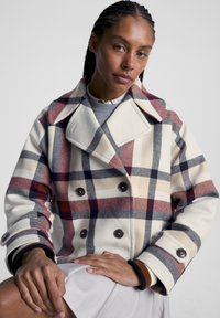 Double-breasted peacoat in cream with red, navy, and beige plaid pattern. Features large buttons and contrasting ribbed cuffs. Soft wool texture.
