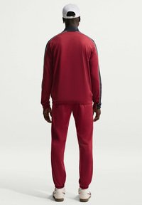 Σετ κόκκινης φόρμας με ναυτικά στοιχεία· το μπουφάν διαθέτει γιακά και φερμουάρ, το παντελόνι έχει ελαστικές μανσέτες. Κατασκευασμένο από απαλό ύφασμα. Φοριέται άσπρο καπέλο.