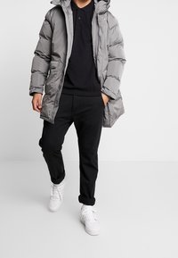 Muž v šedé péřové bundě přes černé polo tričko a černé kalhoty, jde s jednou rukou v kapse a v bílých teniskách.