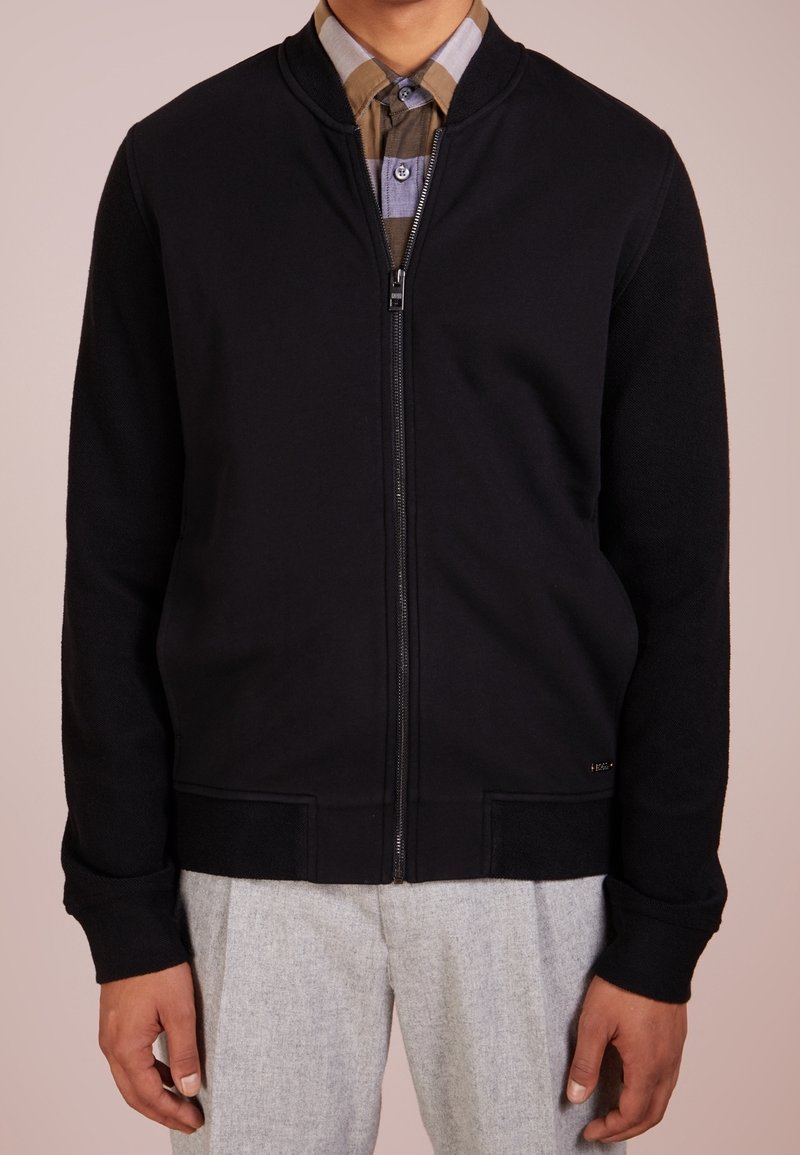Uomo che indossa una giacca nera con zip sopra una camicia a quadri marrone, con pantaloni grigi chiari, su uno sfondo beige.
