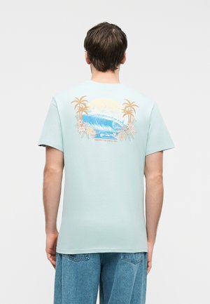 Muž v svetlomodrom tričku s tropickou potlačou oceánskej vlny, paliem a kvetov, oblečený v modrých džínsoch, otočený chrbtom.