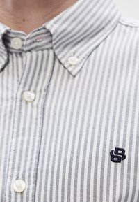 Ριγέ πουκάμισο με κουμπιά σε λευκό και ναυτικό μπλε, με κλασικό γιακά και κέντημα λογότυπου σε ναυτικό μπλ�ε στο αριστερό μέρος. Το ύφασμα φαίνεται υφασμένο.