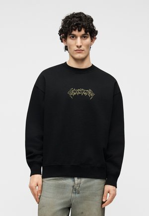 Jeune homme aux cheveux bouclés et foncés portant un sweat-shirt noir avec un petit texte graphique jaune et un jean gris clair, debout devant un fond uni.