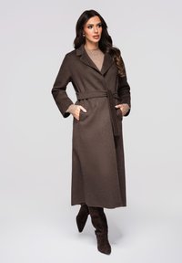 Brun uld trenchcoat med bælte, lange ærmer og krave, med sidelommer, båret over en beige top og brune støvler.