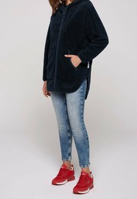 Marinblå fleecejacka med huva, framfickor, dragkedjestängning och sidofickor, ihopparad med ljusblå skinny jeans och röda sneakers.