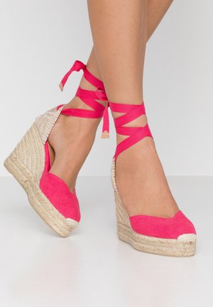 Fødder iført lyserøde kile-espadrillos med ankelbånd og flettede jutesåler på hvid baggrund.