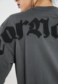 Grå bomulls t-shirt med stor svart broderad text på baksidan. Tyget verkar mjukt och slätt med en avslappnad passform.