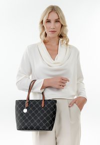 Zwarte tote bag met ruitpatroon, bruine leren handvatten en een zilveren bedel. Draagt met een witte top met col en lichtgekleurde broeken.