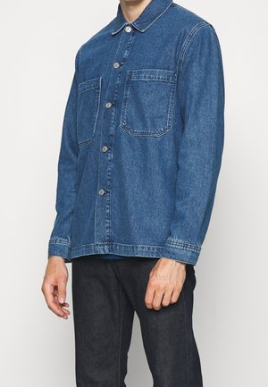 Homme portant une veste en denim bleu foncé boutonnée avec deux poches poitrine et un pantalon noir, debout devant un fond clair uni.