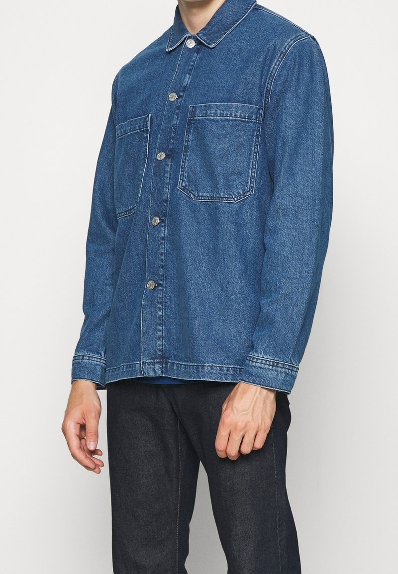 Homme portant une veste en denim bleu foncé boutonnée avec deux poches poitrine et un pantalon noir, debout devant un fond clair uni.