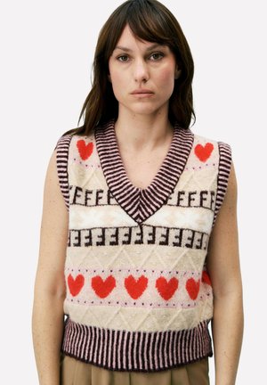 Femme portant un gilet sans manches en maille avec des cœurs rouges, des motifs géométriques et des bordures rayées, debout devant un fond uni.