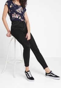 Haut floral marine à manches courtes associé à un jean skinny noir taille haute et des baskets noires. Modèle assis sur un tabouret blanc.