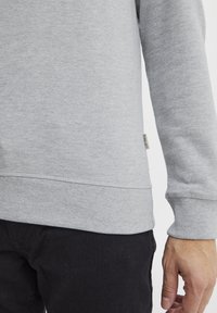 Sudadera gris con puños y dobladillo acanalados, que presenta una pequeña etiqueta de la marca en el lateral. El material parece suave y con textura.