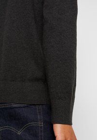Maglione grigio scuro con una texture liscia, caratterizzato da polsini e orlo a coste, abbinato a jeans di denim scuro con dettagli di cucitura visibili.