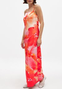 Moteris vilkinti be rankovių, ilga suknele su plonomis petnešomis ir ryškiu raudonų bei oranžinių gėlių raštu, derinama su baltais austais batais.