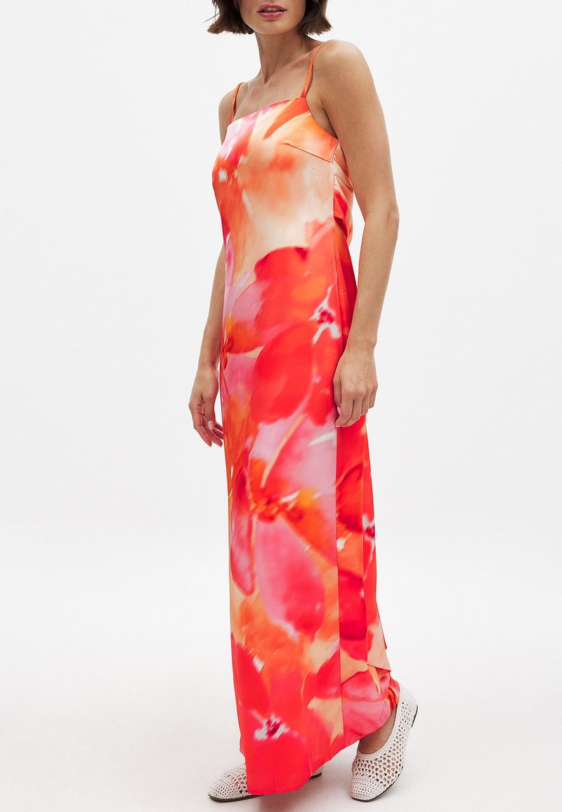 Moteris vilkinti be rankovių, ilga suknele su plonomis petnešomis ir ryškiu raudonų bei oranžinių gėlių raštu, derinama su baltais austais batais.