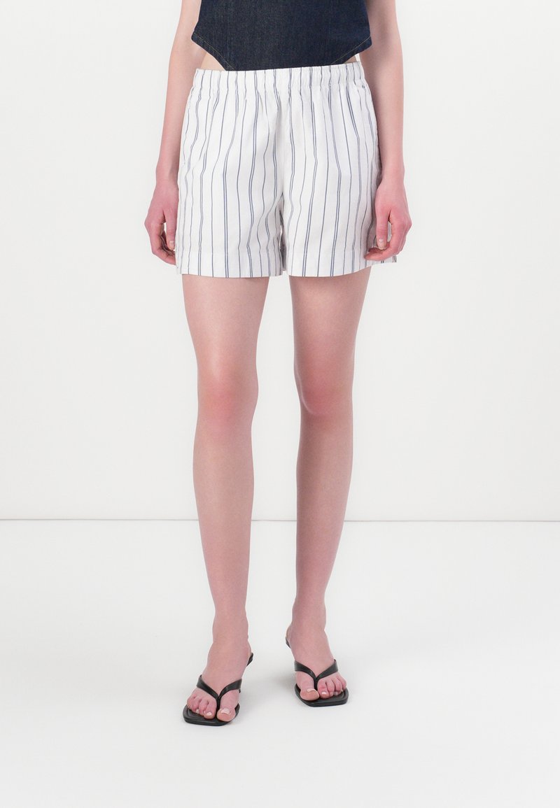 Shorts blancs avec de fines rayures verticales bleues marine, taille élastique et coupe décontractée. Associés à des tongs noires.