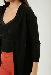 Schwarze Strickjacke mit strukturiertem Zickzackmuster, offener Vorderseite, langen Ärmeln und gerippten Bündchen, kombiniert mit einem schwarzen Oberteil und orangefarbenen Hosen.