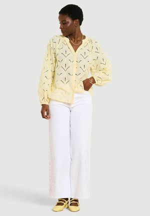 Žena oblečená v žltej vzorovanej blúzke s nafúknutými rukávmi, biele široké nohavice, žlté topánky a zlaté šperky, stojaca pred neutrálnym pozadím.
