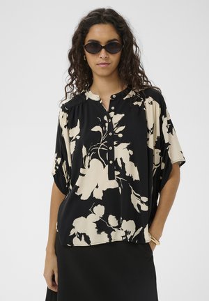 Žena s čiernymi slnečnými okuliarmi a čiernou blúzkou s béžovými kvetinovými vzormi, v kombinácii s čiernou sukňou a zlatým náramkom.